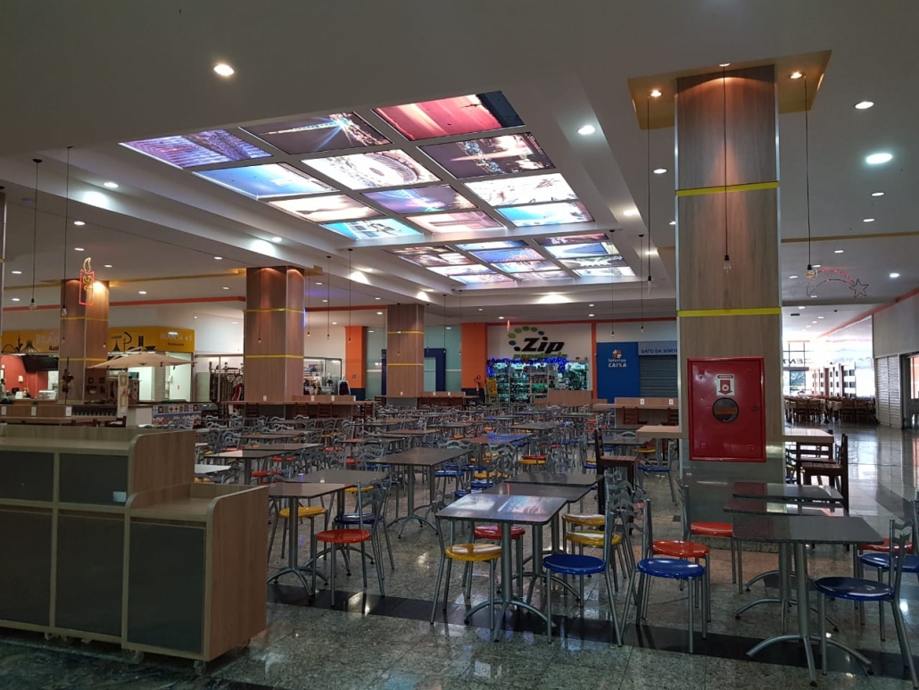 Praça de alimentação do mall vazia, com forro iluminado por painéis retroiluminados e mesas organizadas