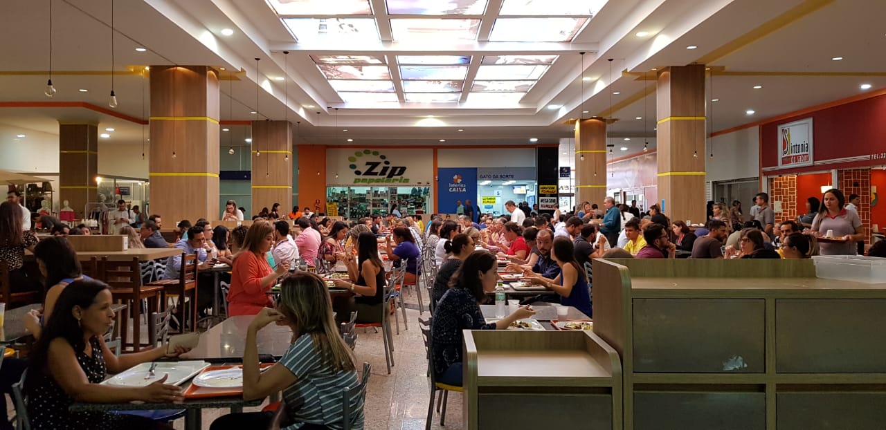 Vista ampla da praça de alimentação do mall, com lojas de serviços ao fundo e público sentado às mesas